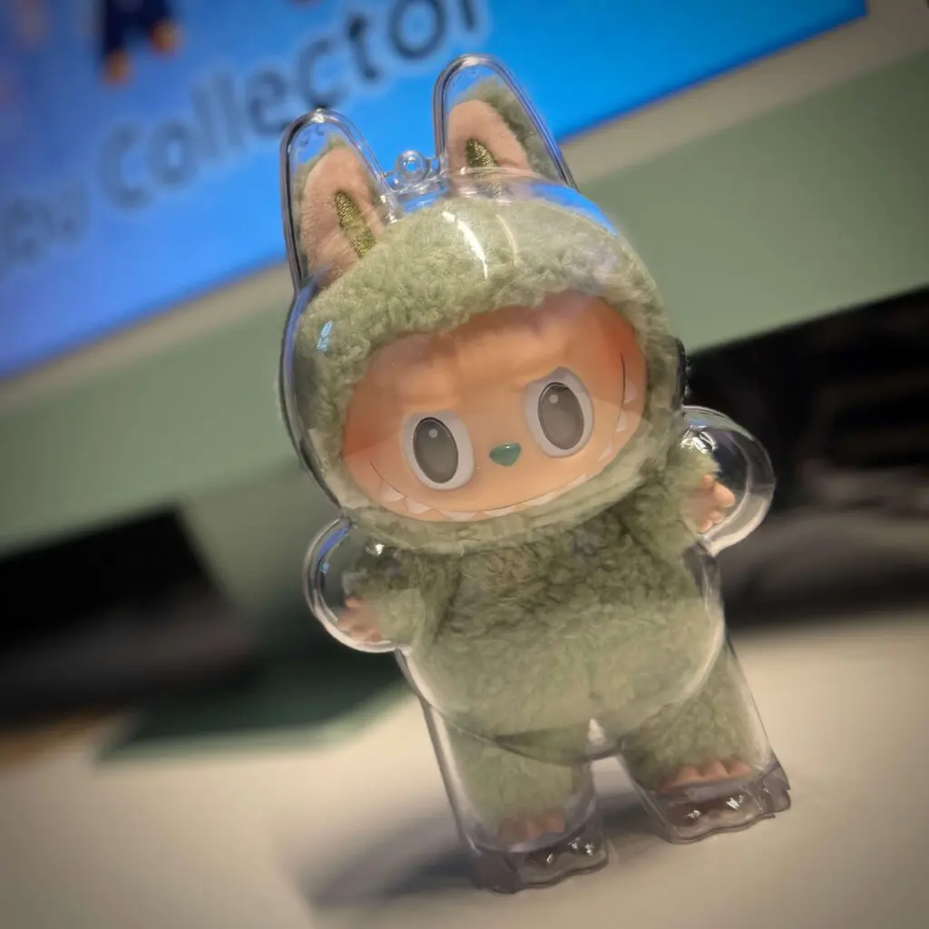 Labubu doll named Green Grape from Exciting Macaron series inside protective plastic case next to iMac