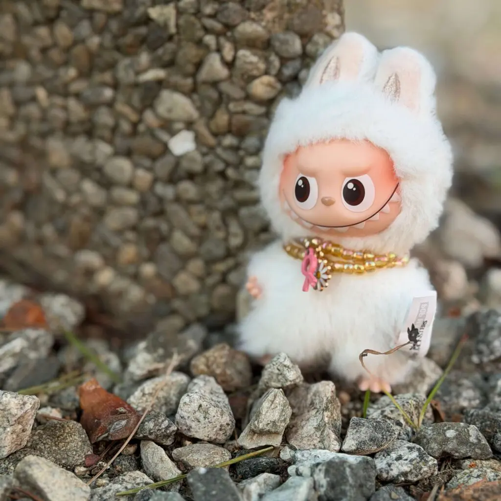 Labubu Pin for Love A Adventure wearing tiny necklace and standing on tiny rocks and next to concrete wall staring right.